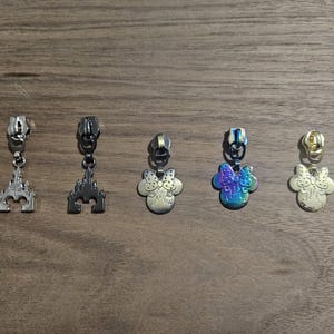 May include: Five metal zipper pulls with decorative charms. Two pulls feature castle designs, while the other three have Minnie Mouse head shapes. The charms are silver, black, iridescent, and gold in color.