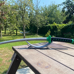 May include: A silver katana sword with a black and green handle is displayed on a silver stand. The sword is resting on a wooden table.