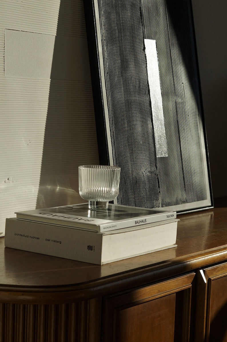 Pode incluir: Uma tigela de vidro transparente e canelado est&aacute; em cima de dois livros, um com a palavra "Bauhaus". Uma obra de arte abstrata em preto e branco emoldurada est&aacute; no fundo. Os objetos est&atilde;o em uma superf&iacute;cie de madeira escura.
