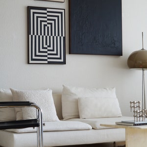 Pu&ograve; includere: Un divano bianco con tre cuscini bianchi, una stampa geometrica in bianco e nero, una stampa astratta in bianco e nero e una stampa beige con piccoli punti. Una sedia in metallo nero si trova di fronte al divano. Un piccolo tavolino da caff&egrave; bianco con un libro e una scultura in metallo si trova di fronte al divano. Un tappeto bianco copre il pavimento.