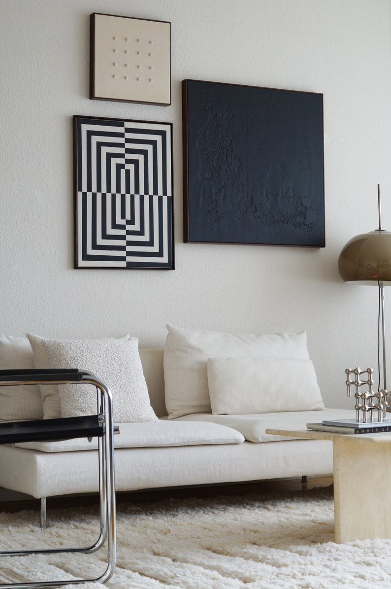 Pu&ograve; includere: Un divano bianco con tre cuscini bianchi, una stampa geometrica in bianco e nero, una stampa astratta in bianco e nero e una stampa beige con piccoli punti. Una sedia in metallo nero si trova di fronte al divano. Un piccolo tavolino da caff&egrave; bianco con un libro e una scultura in metallo si trova di fronte al divano. Un tappeto bianco copre il pavimento.