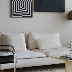 Pu&ograve; includere: Un divano bianco con due cuscini bianchi e un'opera d'arte incorniciata con stampa geometrica in bianco e nero appesa al muro dietro. Una sedia in metallo nero &egrave; in primo piano.