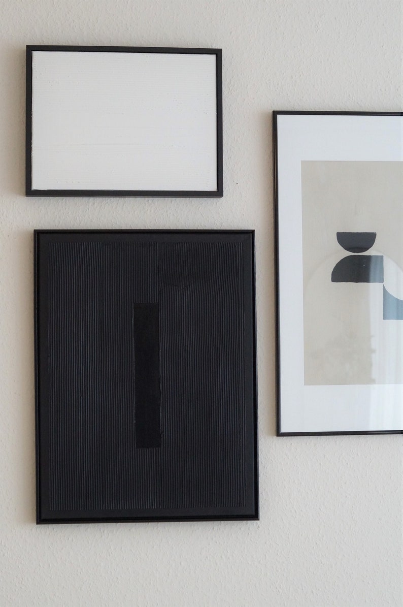 Pode incluir: Tr&ecirc;s obras de arte abstratas emolduradas. A moldura superior cont&eacute;m uma tela branca. A moldura do meio apresenta uma tela preta texturizada com um ret&acirc;ngulo preto vertical. A moldura direita cont&eacute;m uma impress&atilde;o com formas pretas e azuis.