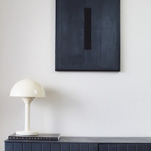 Pode incluir: Cena interior moderna com uma pintura abstrata emoldurada em preto com um ret&acirc;ngulo preto vertical. Uma l&acirc;mpada de mesa branca est&aacute; em um arm&aacute;rio azul escuro, com um livro intitulado "Annie Leibovitz".