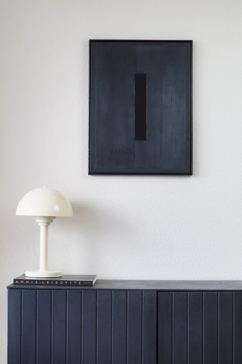 Pode incluir: Cena interior moderna com uma pintura abstrata emoldurada em preto com um ret&acirc;ngulo preto vertical. Uma l&acirc;mpada de mesa branca est&aacute; em um arm&aacute;rio azul escuro, com um livro intitulado "Annie Leibovitz".