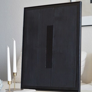 Pode incluir: Obra de arte abstrata emoldurada em preto com uma superf&iacute;cie texturizada e um detalhe retangular. Duas velas brancas em suportes dourados est&atilde;o em frente &agrave; obra de arte. A obra &eacute; exibida em uma superf&iacute;cie de m&aacute;rmore.