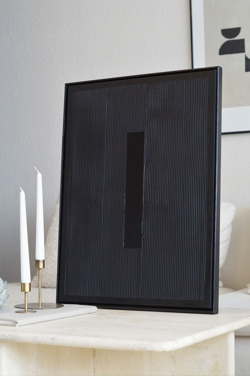 Pode incluir: Obra de arte abstrata emoldurada em preto com uma superf&iacute;cie texturizada e um detalhe retangular. Duas velas brancas em suportes dourados est&atilde;o em frente &agrave; obra de arte. A obra &eacute; exibida em uma superf&iacute;cie de m&aacute;rmore.