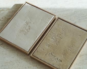 May include: Two framed abstract art pieces. One has a light beige textured surface, the other a darker beige. Both are in light brown wooden frames, set against a light-colored background.