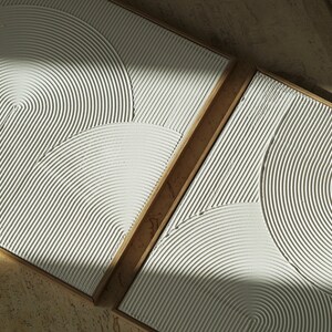 May include: Two square wall art pieces with a raised, textured design of concentric circles. The pieces are framed in natural wood.