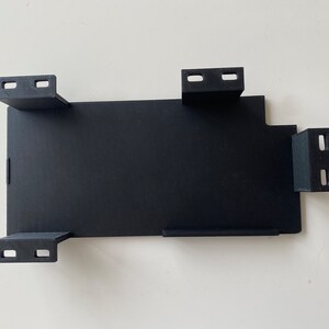 May include: Black plastic bracket with four mounting holes and a rectangular cutout. The bracket is designed for mounting a device or component.