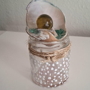 May include: A clear glass jar decorated with small white painted dots and wrapped with natural twine around the neck. Inside the jar sits a large, open oyster shell containing a smooth, translucent amber-colored sphere and small green glass shards.