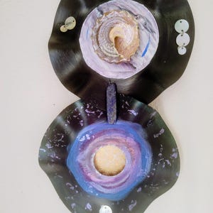 May include: Two black vinyl records with painted designs. The top record has a white, purple, and gold design with a seashell in the center. The bottom record has a blue, purple, and white design with a gold circle in the center. Both records have white sequins around the edges.