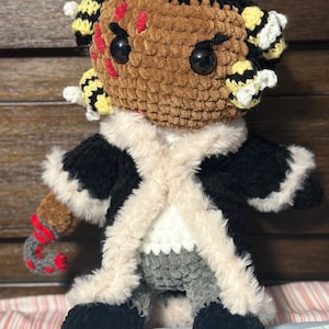 May include: A crocheted plush toy with a brown face, black eyes, and a black and white fur coat. The toy has a red and white candy cane in its hand and yellow and black bee-shaped decorations on its head.