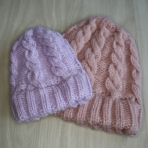 Pode incluir: Dois chapéus de malha, um em cor rosa claro e o outro em cor pêssego pálido. Ambos os chapéus têm um padrão de malha de cabo.