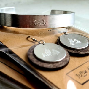 May include: A silver-toned cuff bracelet and matching earrings. The bracelet and earrings feature a forest design. The earrings have a wooden base with a silver disc on top. The jewelry is packaged in a clear plastic bag.