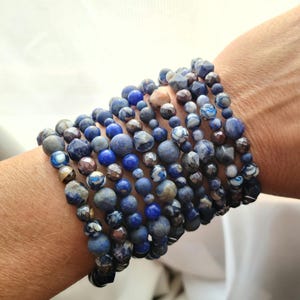 May include: A stack of blue and gray beaded bracelets on a person's wrist. The bracelets are made of various types of stones, including lapis lazuli, agate, and jasper.