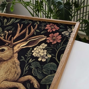 May include: A framed print of a stylized rabbit with antlers, surrounded by a dark floral pattern with pink and white flowers. The frame is made of natural wood.