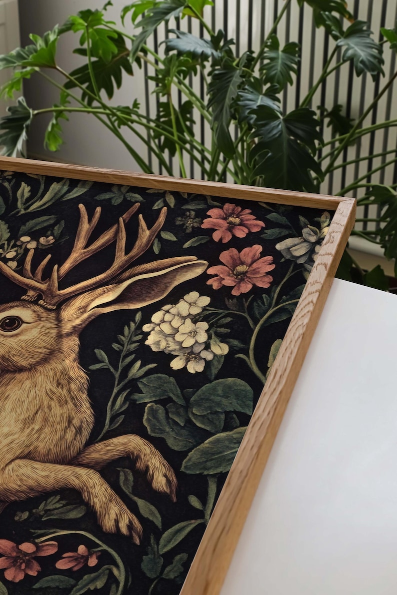 May include: A framed print of a stylized rabbit with antlers, surrounded by a dark floral pattern with pink and white flowers. The frame is made of natural wood.