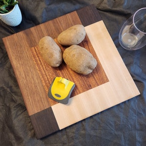 Puede incluir: Una tabla de cortar de madera con un centro de madera clara y un borde de madera oscura. Hay tres patatas en la tabla y un raspador amarillo y gris.