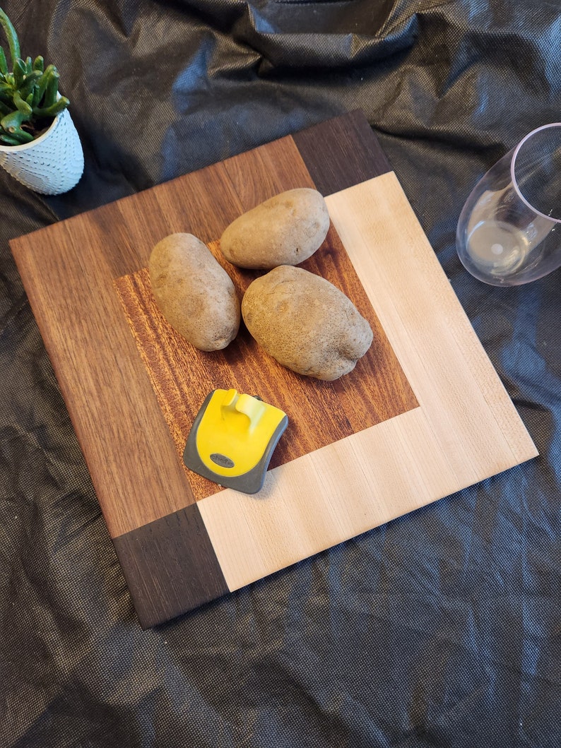 Puede incluir: Una tabla de cortar de madera con un centro de madera clara y un borde de madera oscura. Hay tres patatas en la tabla y un raspador amarillo y gris.