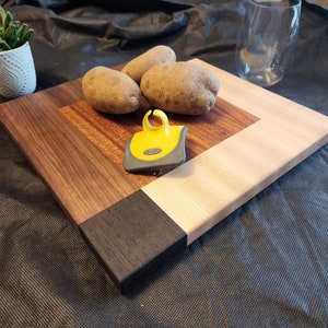 Puede incluir: Una tabla de cortar de madera con un patr&oacute;n de veta de madera marr&oacute;n claro, marr&oacute;n oscuro y negro. Tres patatas marrones y un pelador de patatas amarillo est&aacute;n sobre la tabla.