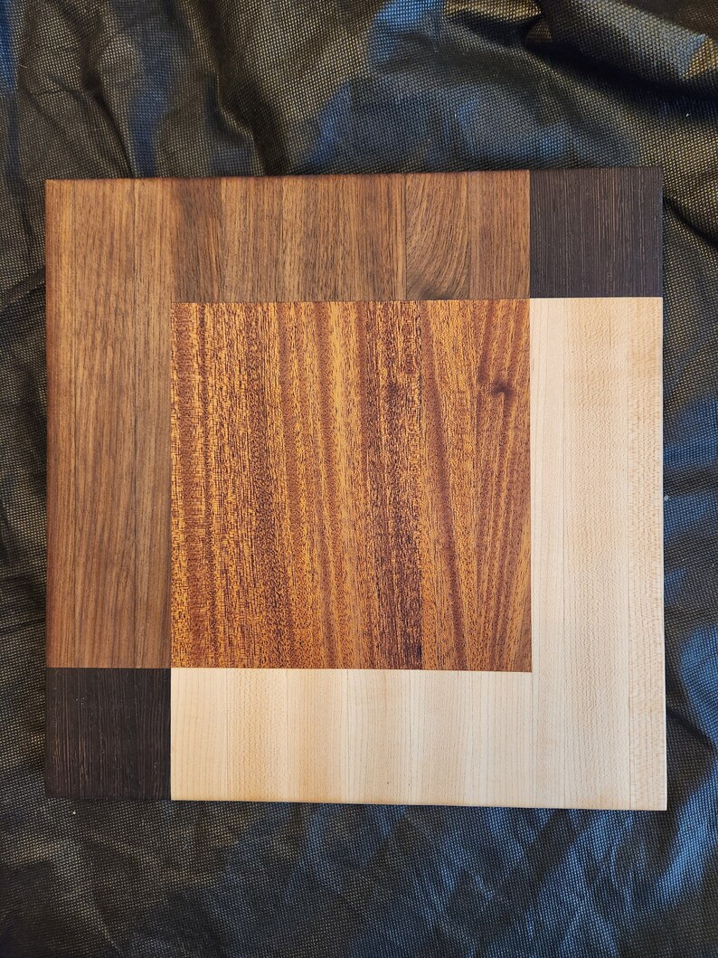 Puede incluir: Una tabla de cortar de madera con un dise&ntilde;o &uacute;nico. La tabla est&aacute; hecha de tres tipos diferentes de madera: nogal, arce y cerezo. El nogal es de color marr&oacute;n oscuro, el arce es de color marr&oacute;n claro y el cerezo es de color marr&oacute;n rojizo.