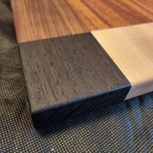 Puede incluir: Primer plano de una tabla de cortar de madera con diferentes tipos de madera, incluyendo una secci&oacute;n de madera oscura te&ntilde;ida. La tabla est&aacute; sobre una superficie negra texturizada.