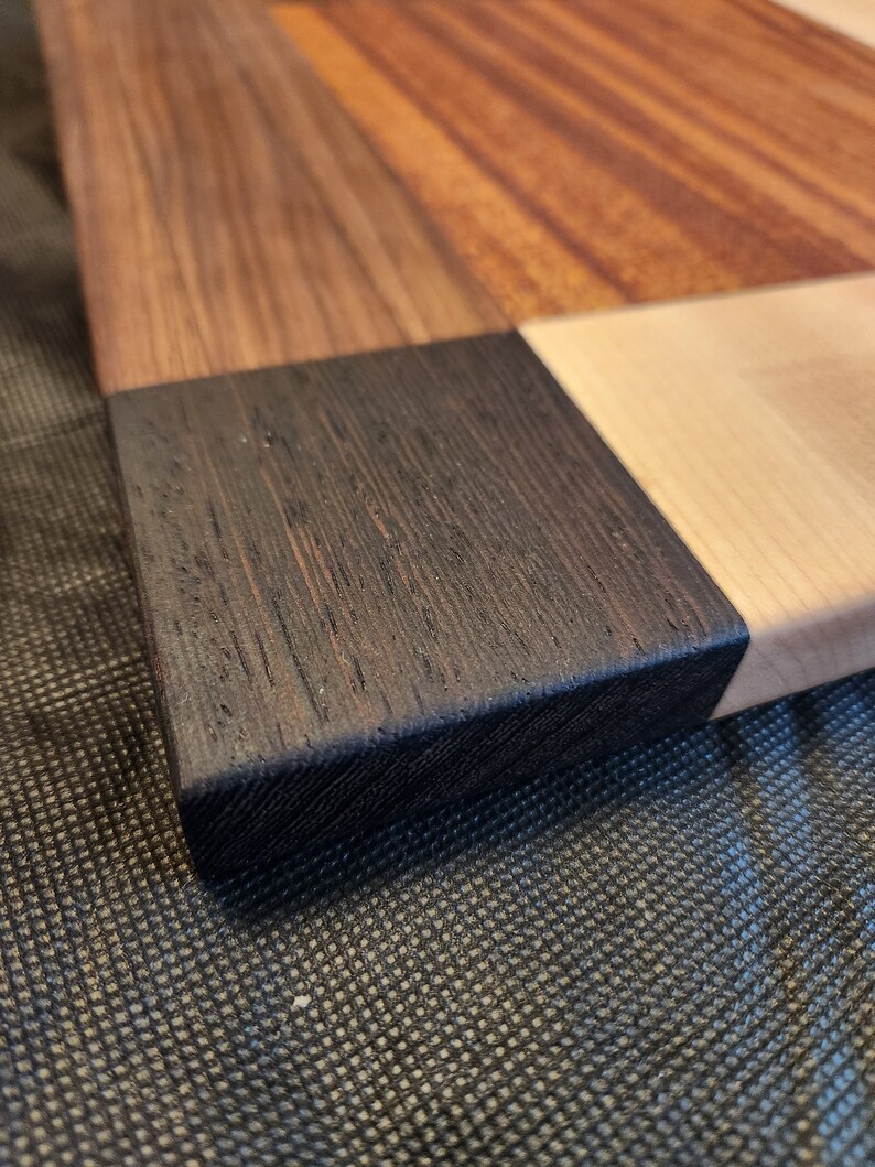 Puede incluir: Primer plano de una tabla de cortar de madera con diferentes tipos de madera, incluyendo una secci&oacute;n de madera oscura te&ntilde;ida. La tabla est&aacute; sobre una superficie negra texturizada.