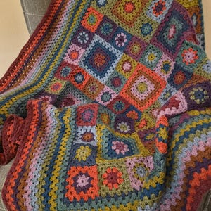 May include: A handmade crochet blanket featuring a multi-colored granny square pattern. The design incorporates various shades of deep red, forest green, navy blue, mustard yellow, and soft lavender, arranged in a repeating geometric square motif.
