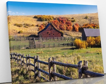 May include: A red wooden barn with a fence in front of it. The barn is surrounded by a field of green grass and trees with fall foliage. The sky is blue and there are clouds in the distance.