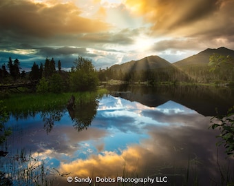 May include: A scenic landscape photograph of a lake reflecting a cloudy sky with a golden sunset. The lake is surrounded by trees and mountains. The sun's rays are visible through the clouds.