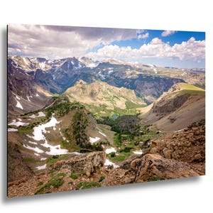 May include: A scenic landscape photograph of a mountain range with snow-capped peaks under a cloudy sky. The foreground features rocky terrain, while a valley with a small lake and patches of snow is visible in the distance.