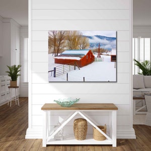 May include: A red barn with a white roof sits in a snowy field with a fence in front of it. The barn is surrounded by trees and a mountain range in the background.