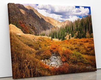 May include: A scenic landscape photograph featuring a mountain range with a valley in the foreground. The valley is covered in a mix of green and yellow foliage, with a small stream running through it. The mountains are covered in trees and have a rocky, brown appearance. The sky is blue with white clouds.