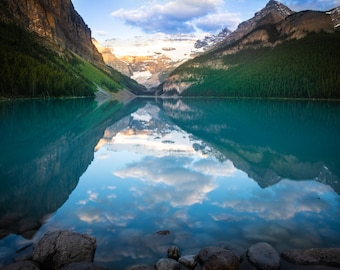 May include: A breathtaking reflection of snow-capped mountains and clouds in a turquoise lake. The water is still and clear, reflecting the sky and the surrounding landscape. The mountains are tall and majestic, with a hint of green trees at their base.