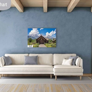 May include: A white couch with pillows sits in a living room with a wooden door and a light blue wall. A framed photograph of a wooden chapel with a mountain backdrop hangs on the wall. The photo is labeled "2x3".