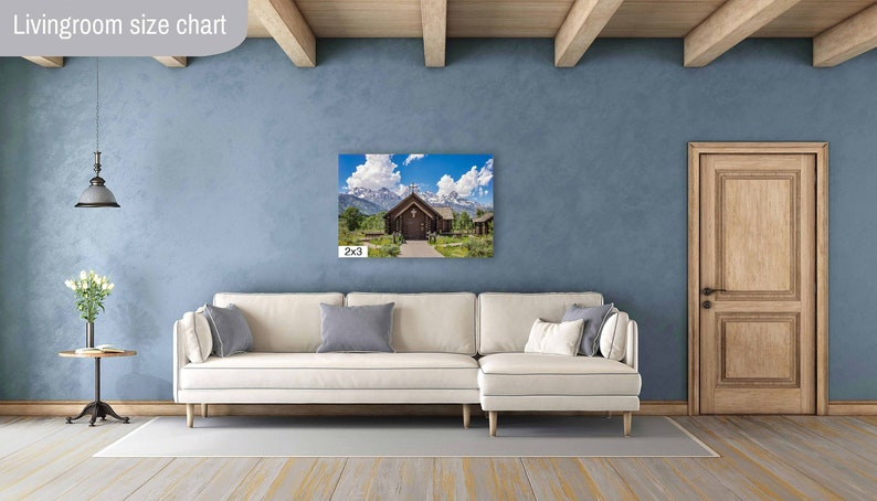 May include: A white couch with pillows sits in a living room with a wooden door and a light blue wall. A framed photograph of a wooden chapel with a mountain backdrop hangs on the wall. The photo is labeled "2x3".