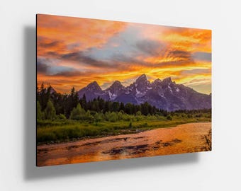 Op de afbeelding: Een landschapsfoto met een bergketen en een rivier op de voorgrond. De lucht is fel oranje en geel, met wolken die in het water worden weerspiegeld. De bergen zijn paars en groen, met besneeuwde toppen.
