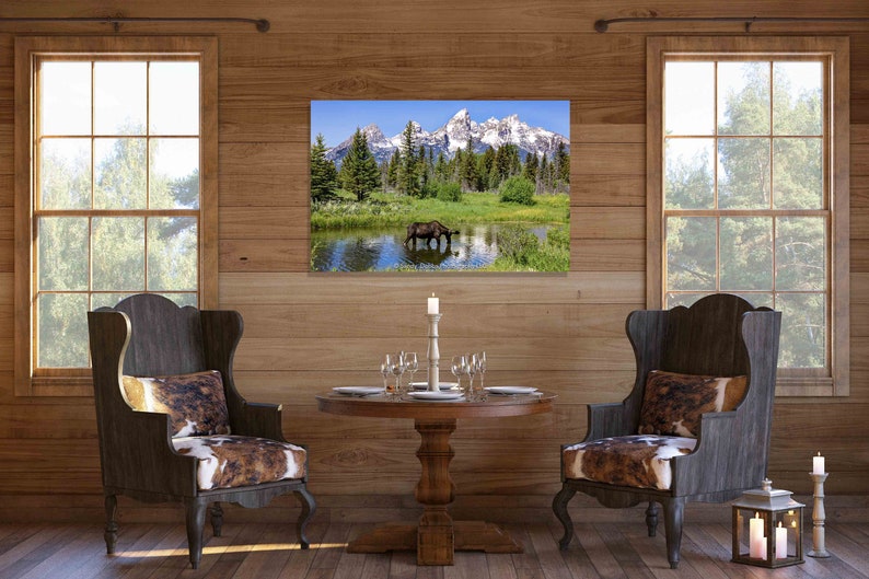 Op de afbeelding: Een rustieke eetkamer met twee stoelen, een ronde houten tafel en een ingelijst afdruk van een eland die drinkt uit een meer voor besneeuwde bergen.