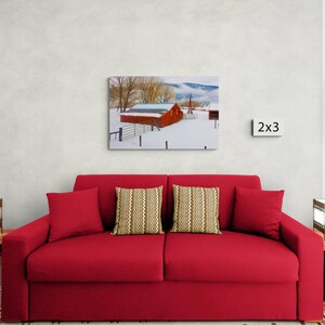 May include: A red barn with a snow-covered roof sits in a snowy field with trees in the background. The barn is surrounded by a fence. The image is framed and displayed on a wall above a red couch with two pillows.