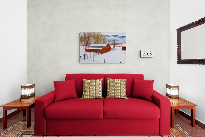 May include: A red barn with a snow-covered roof sits in a snowy field with trees in the background. The barn is surrounded by a fence. The image is framed and displayed on a wall above a red couch with two pillows.