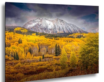 Puede incluir: Impresión de un paisaje montañoso con picos nevados bajo un cielo tormentoso. El primer plano está lleno de árboles amarillos vibrantes, lo que sugiere una escena otoñal. Los árboles de hoja perenne de color verde oscuro salpican el paisaje.