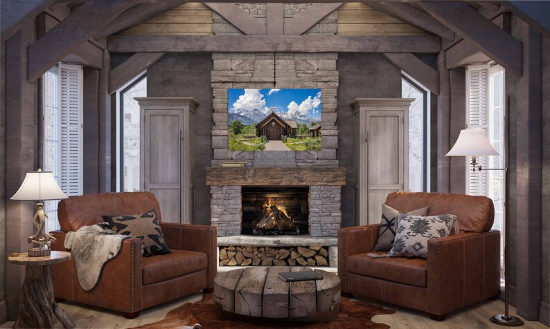 May include: A rustic living room with a stone fireplace, two brown leather armchairs, a wooden coffee table, and a framed landscape photograph of a wooden church in a mountain valley.