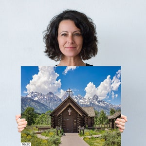 May include: A photograph of a wooden chapel with a cross on the roof, set against a backdrop of snow-capped mountains and a bright blue sky with fluffy white clouds. The chapel has a wooden walkway leading to the entrance.
