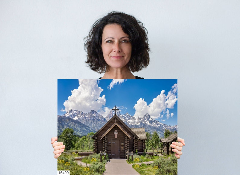May include: A photograph of a wooden chapel with a cross on the roof, set against a backdrop of snow-capped mountains and a bright blue sky with fluffy white clouds. The chapel has a wooden walkway leading to the entrance.