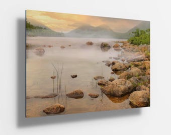 Puede incluir: Una fotografía de paisaje que representa un lago con montañas en el fondo. El lago está tranquilo e inmóvil, con una ligera niebla que se eleva del agua. Las montañas están cubiertas de árboles y se recortan contra un cielo nublado. Los colores de la fotografía son suaves y apagados, creando una atmósfera pacífica y serena.