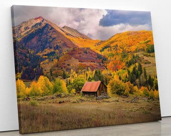 May include: A canvas print of a mountain landscape in autumn. The scene features a rustic wooden cabin with a red roof nestled in a valley. The surrounding hills are covered in vibrant yellow, orange, and red foliage, with a cloudy sky overhead.