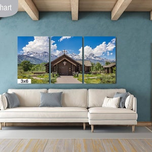 May include: A three-panel canvas print of a wooden cabin with a cross on the roof, set against a backdrop of snow-capped mountains and a blue sky with white clouds. The print is displayed on a white couch in a living room with a light blue wall and wooden beams.