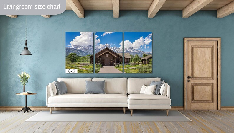 May include: A three-panel canvas print of a wooden cabin with a cross on the roof, set against a backdrop of snow-capped mountains and a blue sky with white clouds. The print is displayed on a white couch in a living room with a light blue wall and wooden beams.