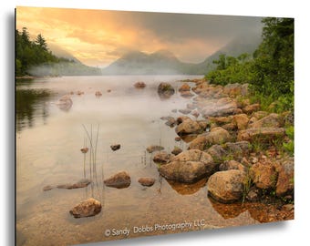 May include: A landscape photograph of a calm lake at sunset with golden light reflecting on the water. The shoreline is lined with numerous brown rocks and green vegetation. Mist rises from the water surface near distant mountains. Sandy Dobbs Photography LLC.